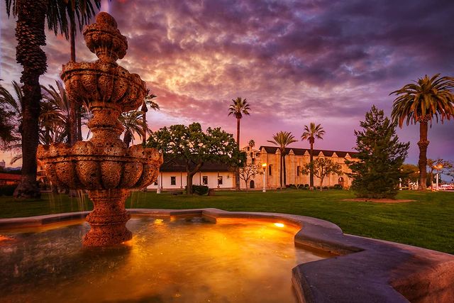 A Quinceanera image of Mission San Gabriel Arc&aacute;ngel with a fountain in front of a building and palm trees